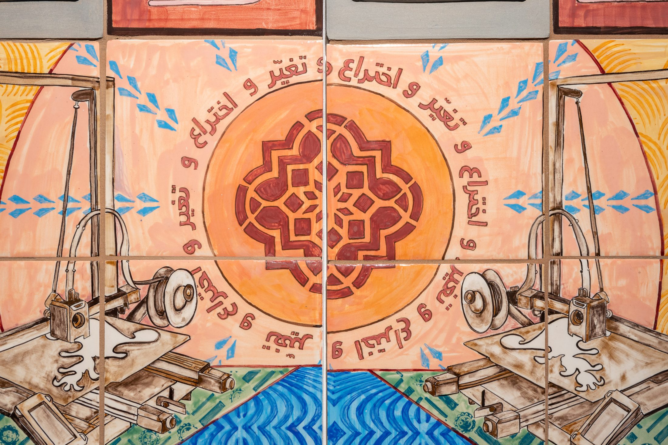 Colourful tiles featuring two industrial machines facing each other, ornate orange geometric pattern in the centre, and Arabic script encircling the pattern.