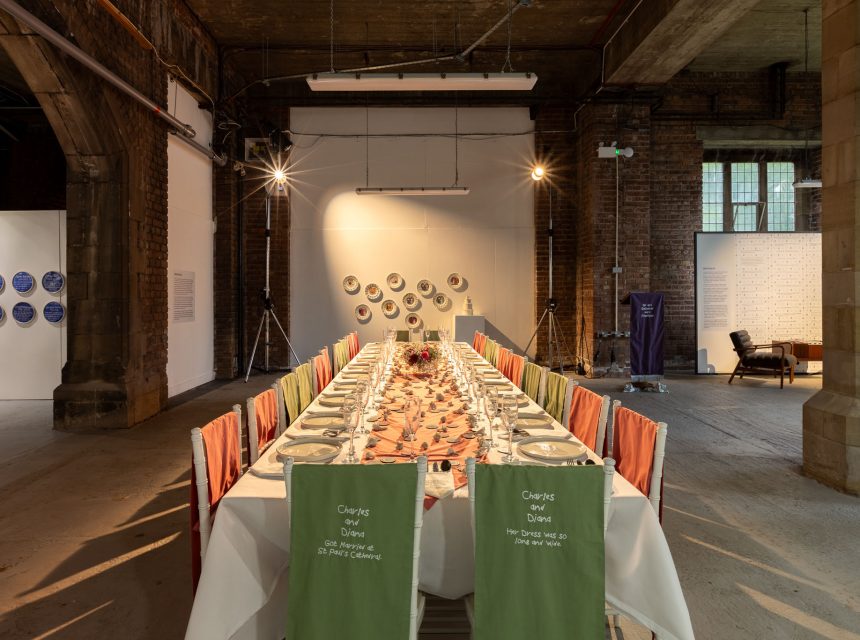 A long, rectangular dining table set for a meal stands in an industrial-style room with exposed brick and stone arches. Colourful fabric covers the chairs, and decorative plates hang on the far wall as part of an exhibition.