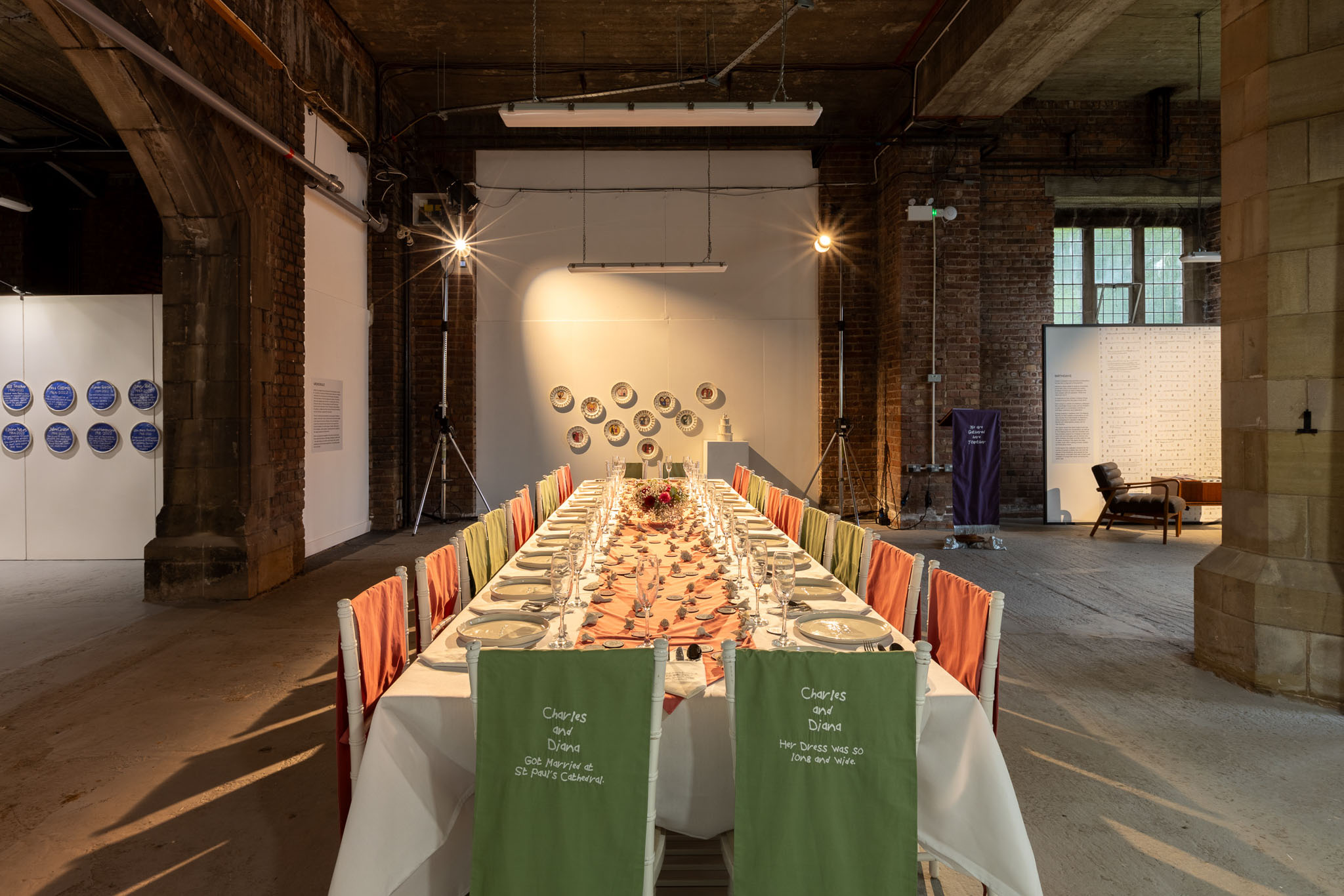 A long, rectangular dining table set for a meal stands in an industrial-style room with exposed brick and stone arches. Colourful fabric covers the chairs, and decorative plates hang on the far wall as part of an exhibition.