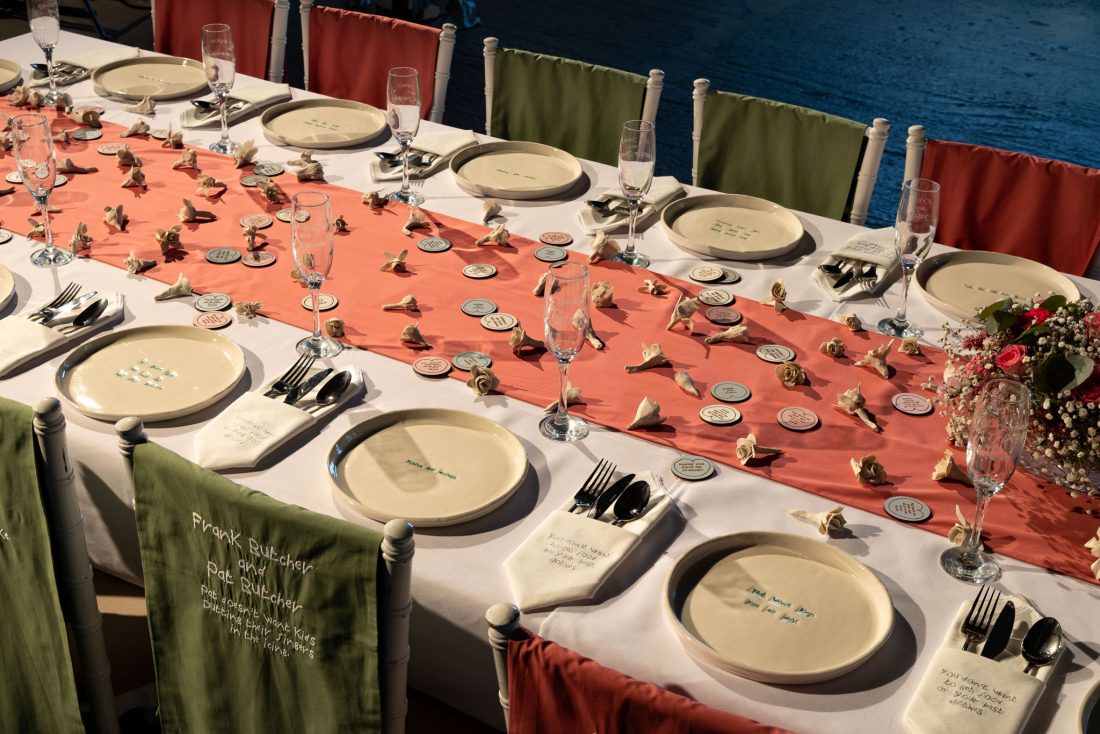 A long table set for a formal event with beige plates, cutlery, champagne glasses, and peach and green table runners, decorated with small paper flowers and a bouquet of gypsophila.