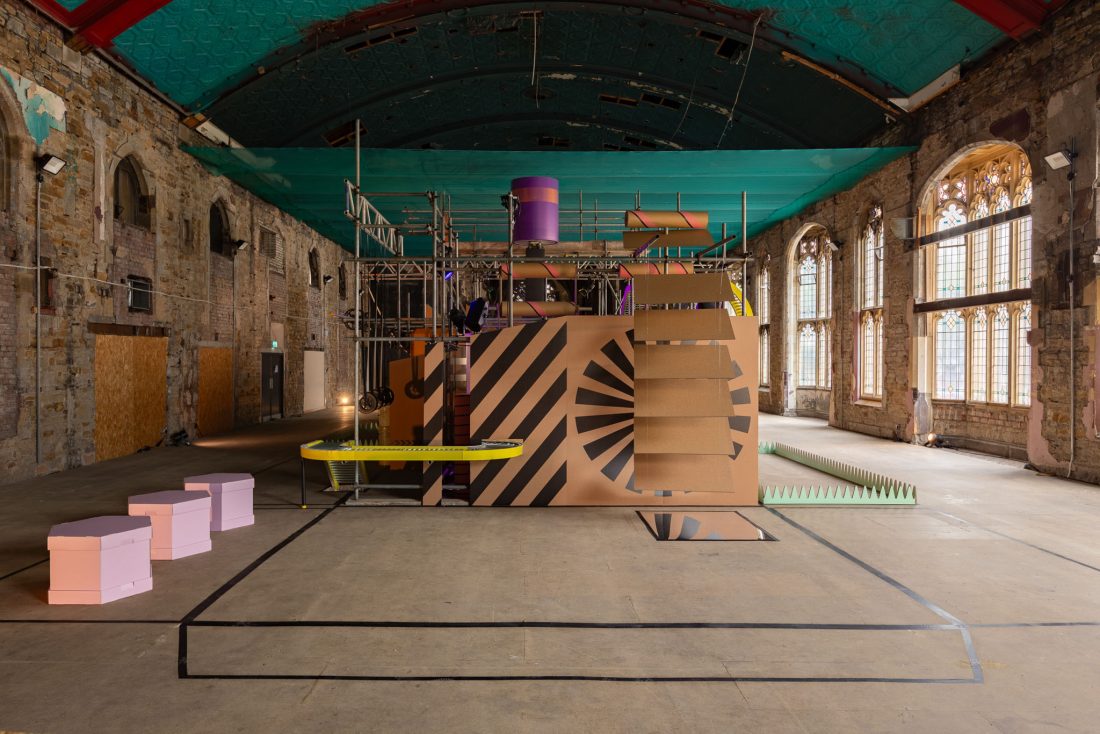 A colourful geometric art installation with stripes, shapes, and scaffolding stands in a large, empty hall with arched windows and old stone walls. Pink, yellow, and green accents are present throughout the scene.