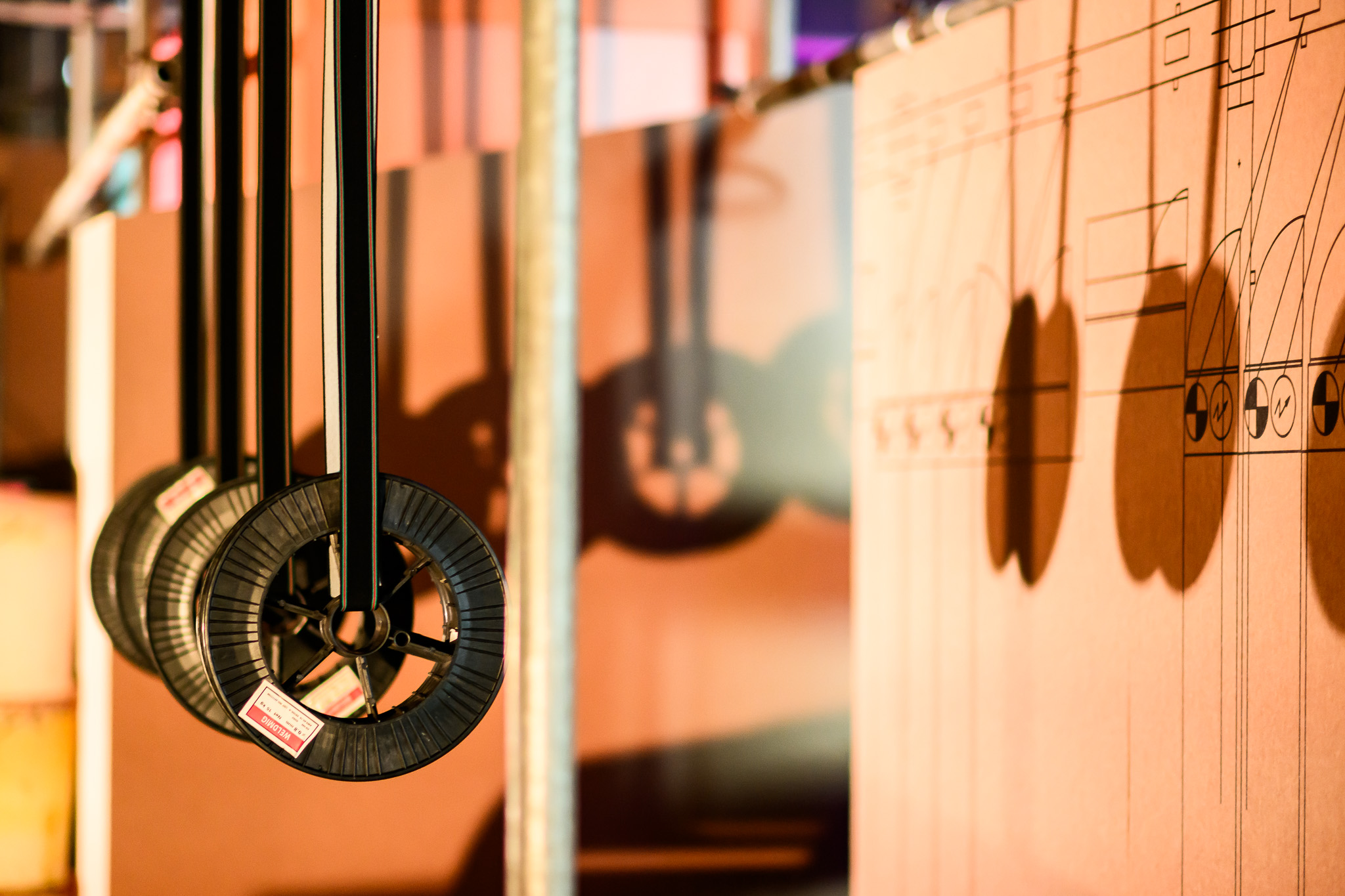 Three black wheels with spokes hang from rods, casting shadows on a cardboard wall with technical drawings. The lighting creates distinct shadows, highlighting the industrial and mechanical theme.