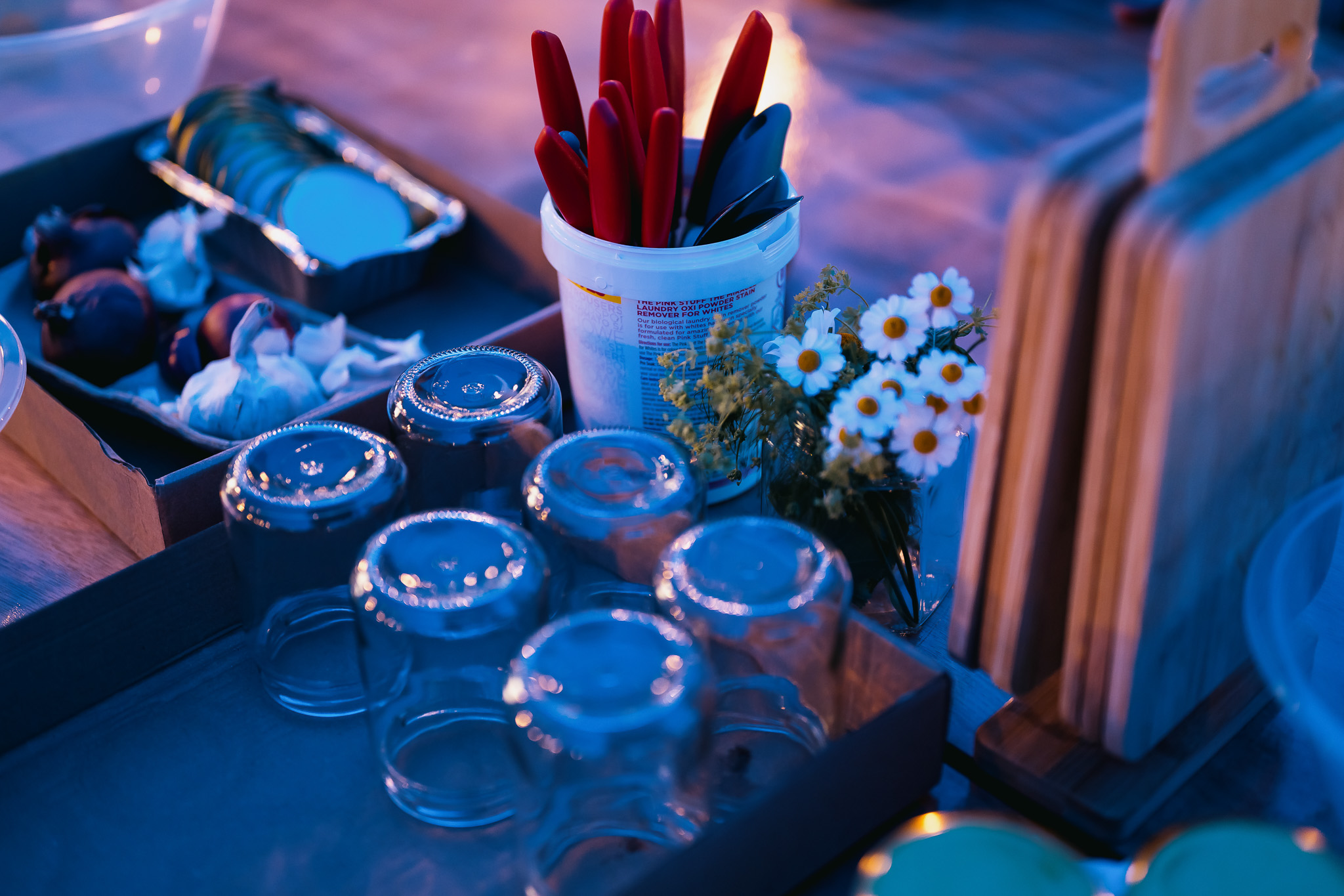 A kitchen setting with clear glasses, a bucket of red knives, white daisies, cutting boards, and assorted kitchen items, bathed in warm blue light.