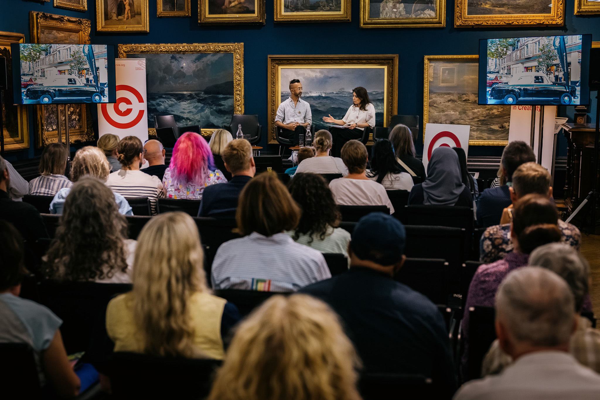 A seated audience listens to Hetain Patel and Mariam Zulfiqar speaking onstage in an art gallery, surrounded by paintings and two large screens displaying outdoor scenes.