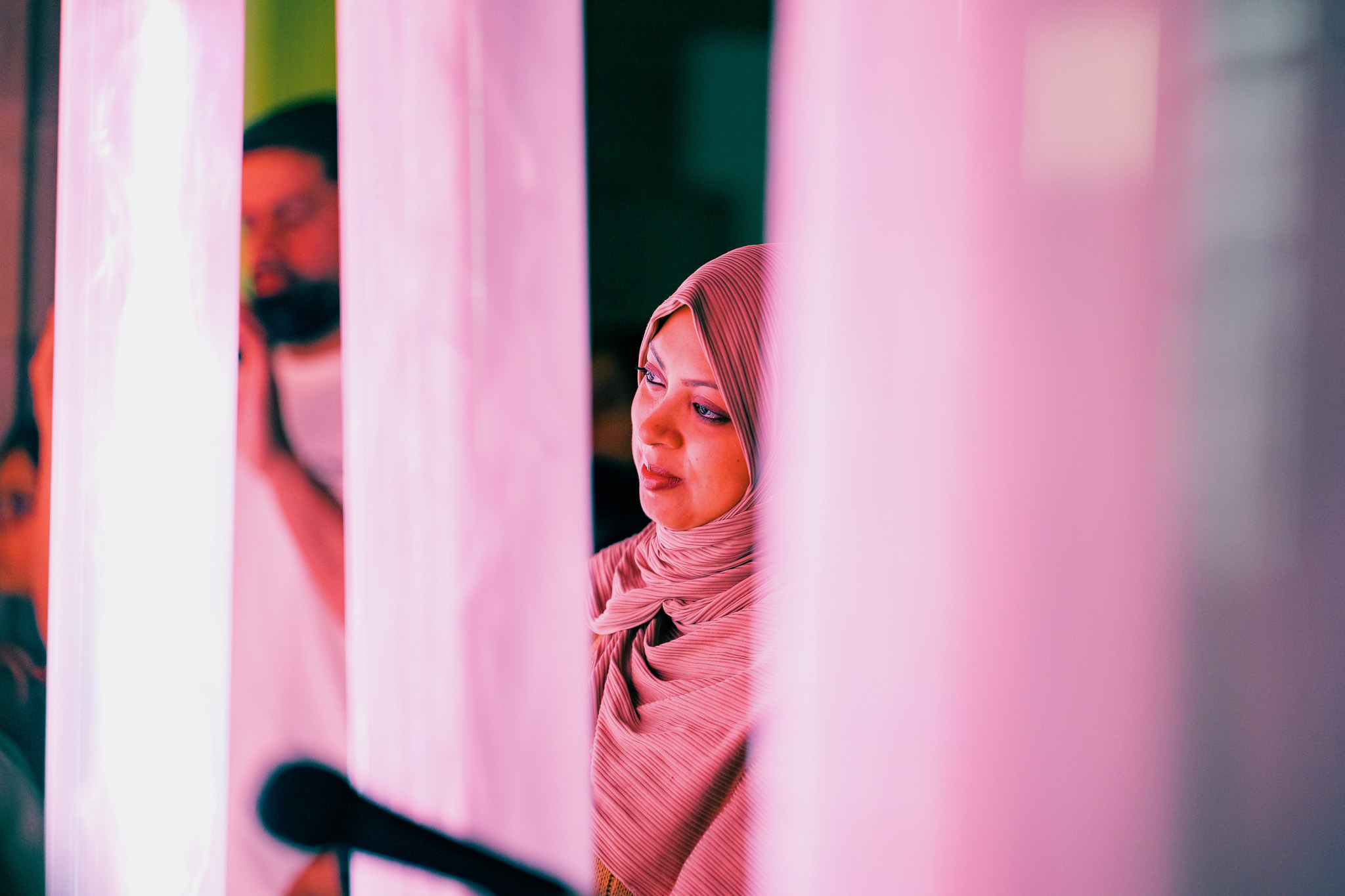 A woman wearing a hijab stands between illuminated pink pipes, softly lit. She gazes to the side, with a microphone visible in the foreground and a blurred figure in the background.