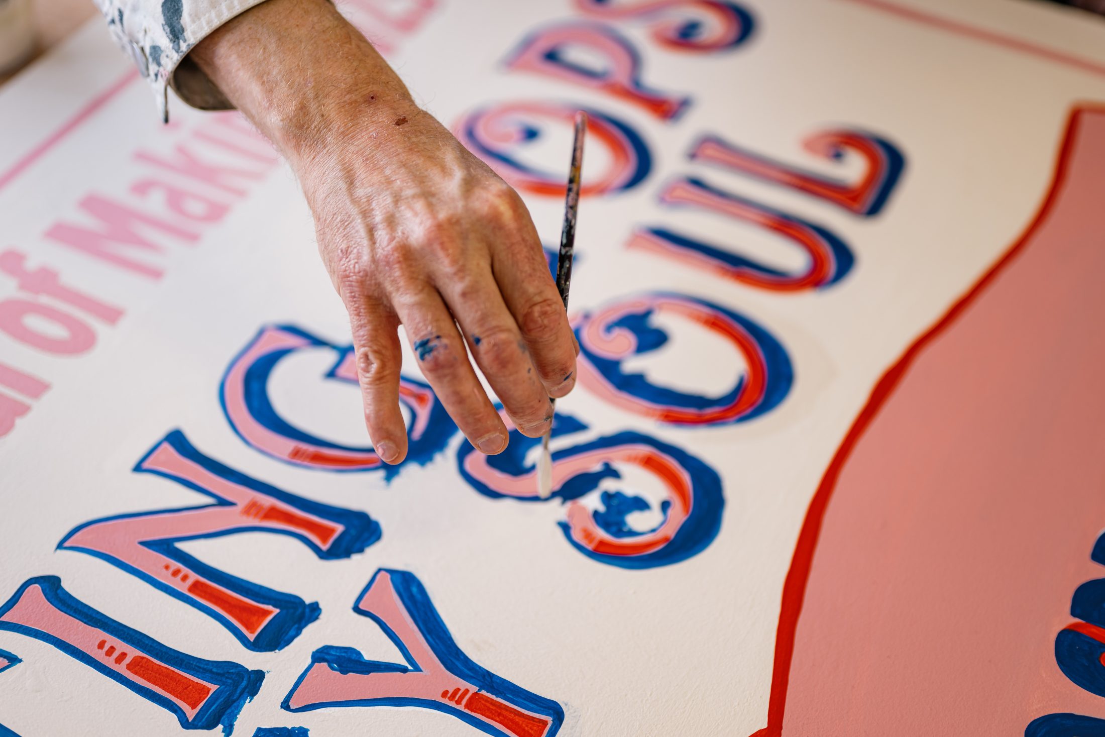 A person carefully paints bold blue and red letters on a white surface, adding detail to the word “SOUL” on a colourful hand-painted sign.