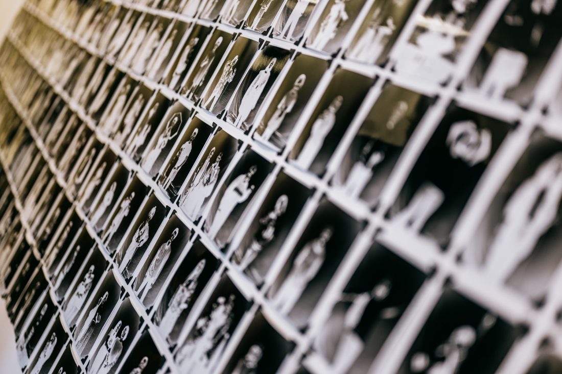 A wall covered in rows of black-and-white portrait photographs, each featuring a person, arranged in a grid pattern and viewed from an angle, creating a sense of depth and repetition.