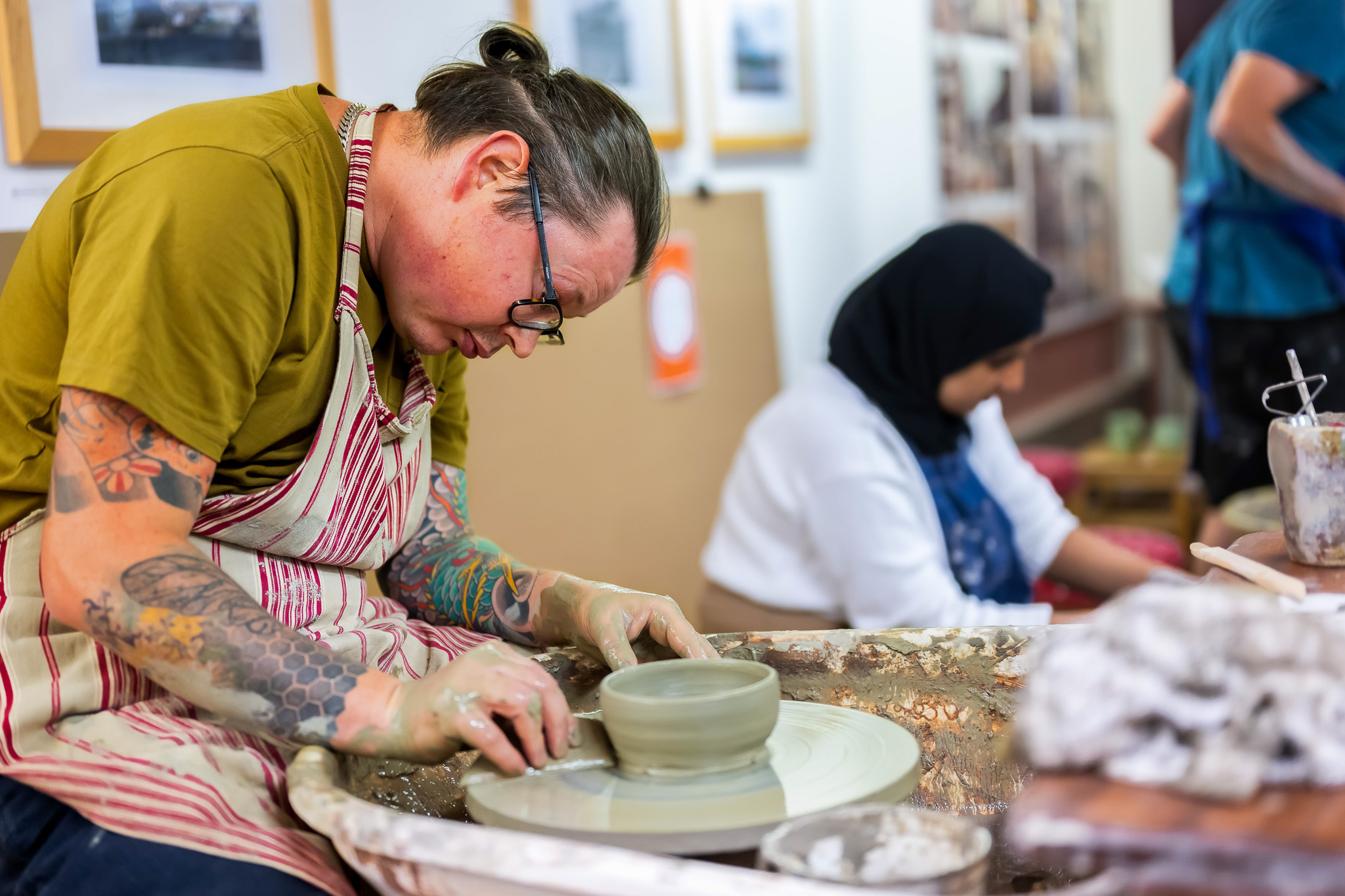 Two people engaged in pottery-making. In the foreground, one person is focused on shaping clay on a pottery wheel, while another works in the background.