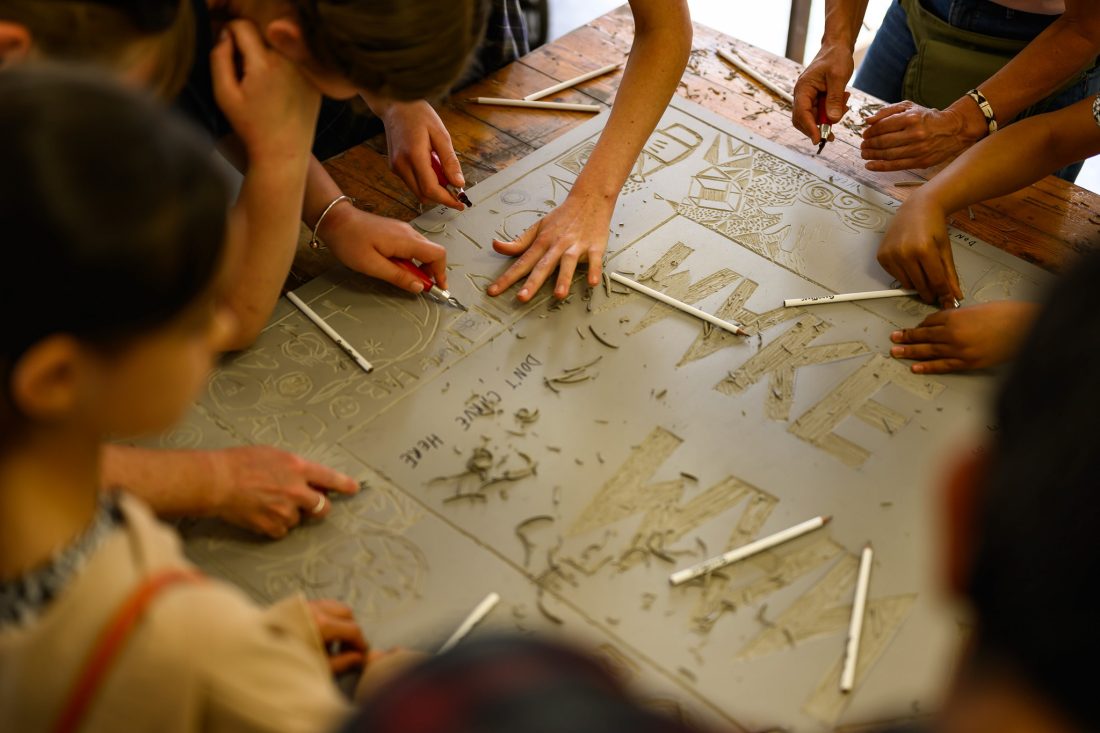 A group of people collaboratively carve a large linoleum sheet at a workshop. Hands are shown holding carving tools, surrounded by shavings and designs.
