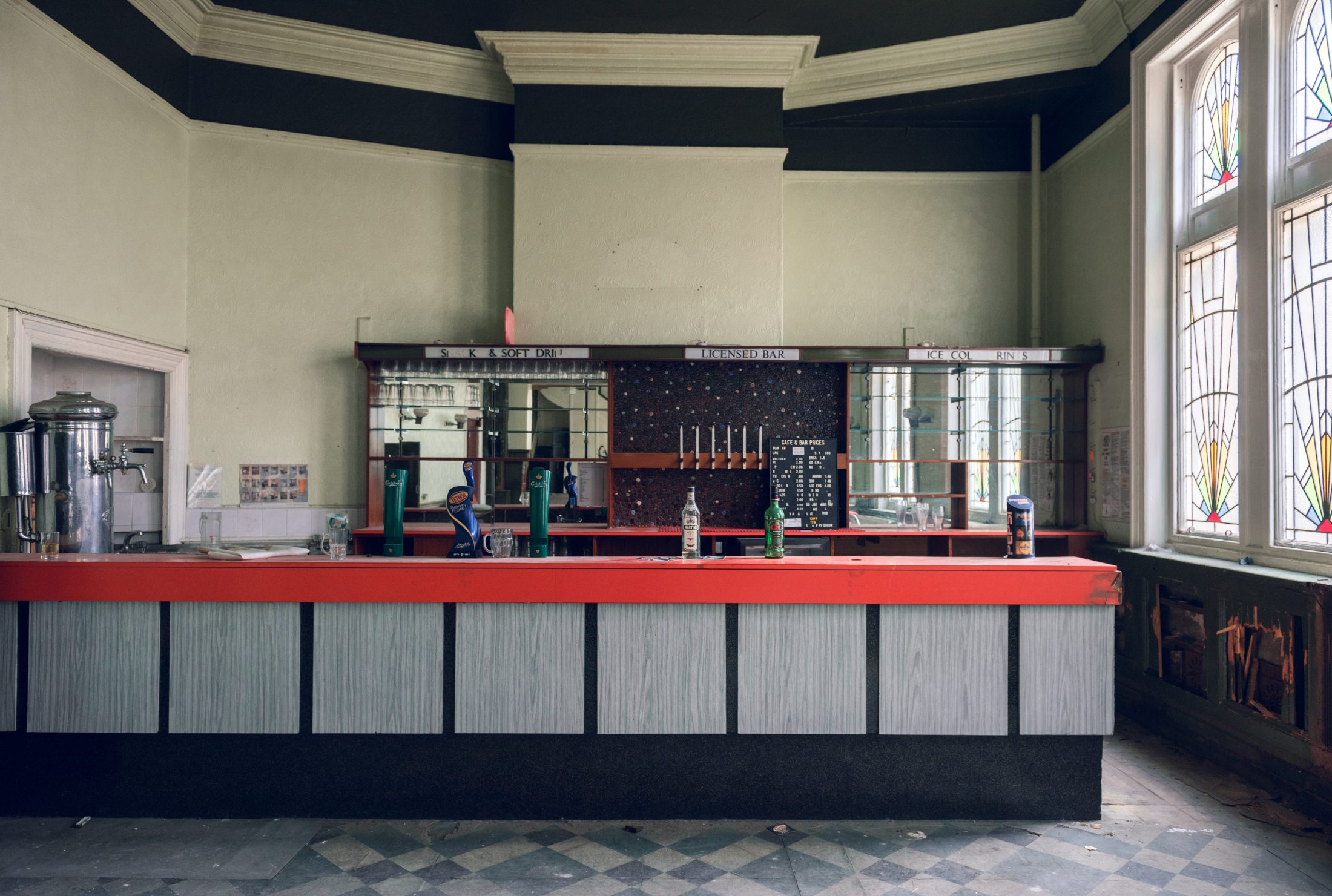 A vintage bar with a red and gray counter, empty glass bottles, and taps. Stained glass windows cast soft light.