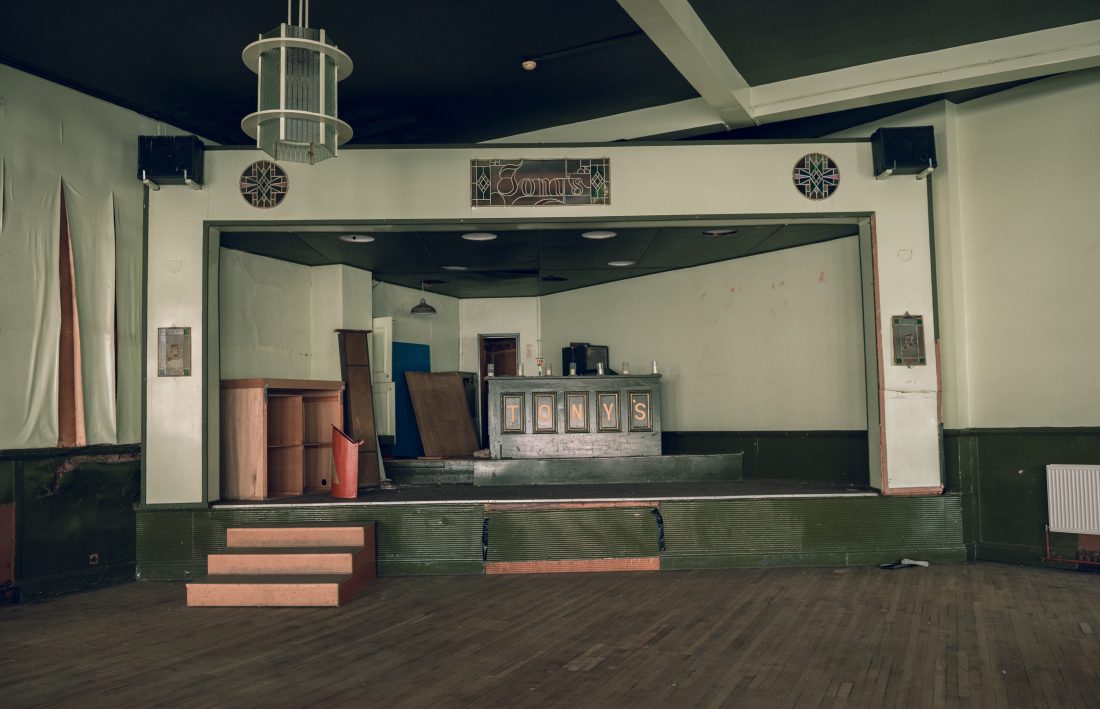 An abandoned, shabby stage with peeling paint and a vintage "Tony's" sign. Empty bar area behind.