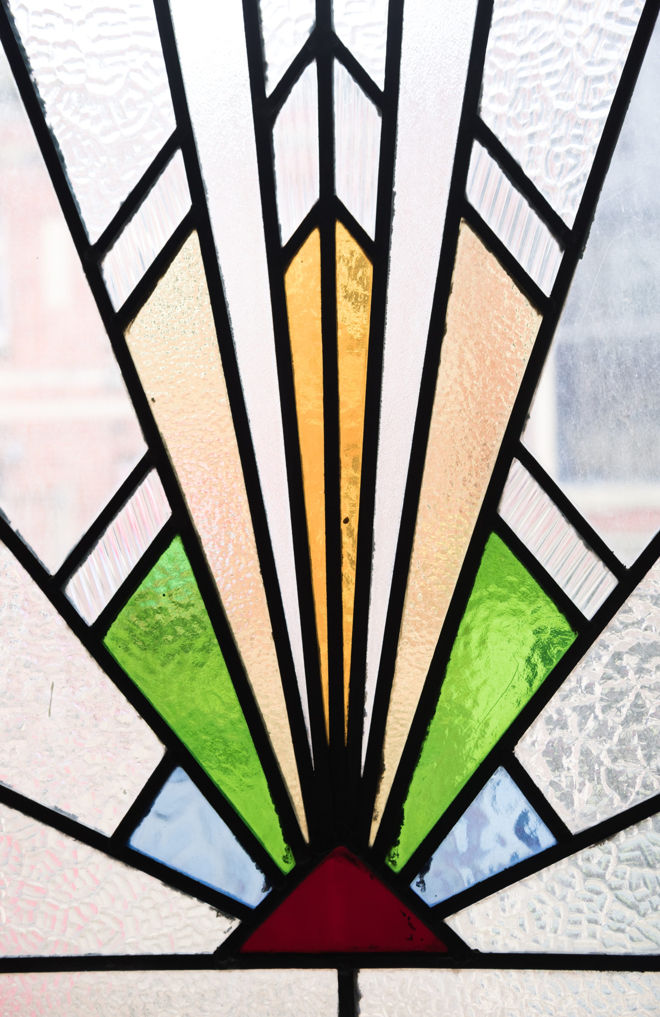 Art Deco stained glass window featuring geometric shapes with bold lines. Central chevron design in gold, green, and blue.