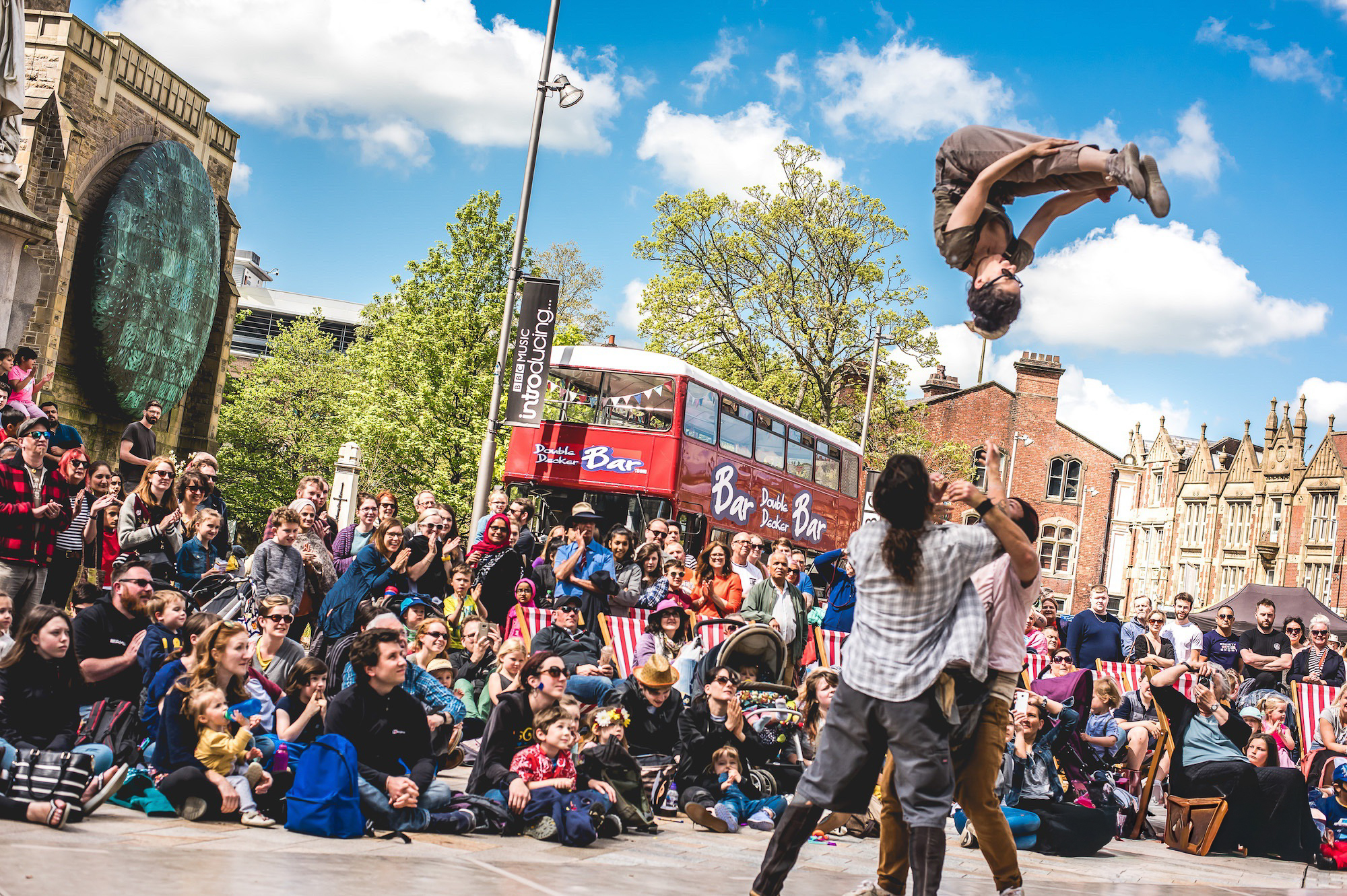 Two performers entertain a large outdoor crowd; one acrobat flips high in the air above the other’s arms. Spectators watch, some seated and some standing, in front of old buildings and a red double-decker bus under a blue sky.