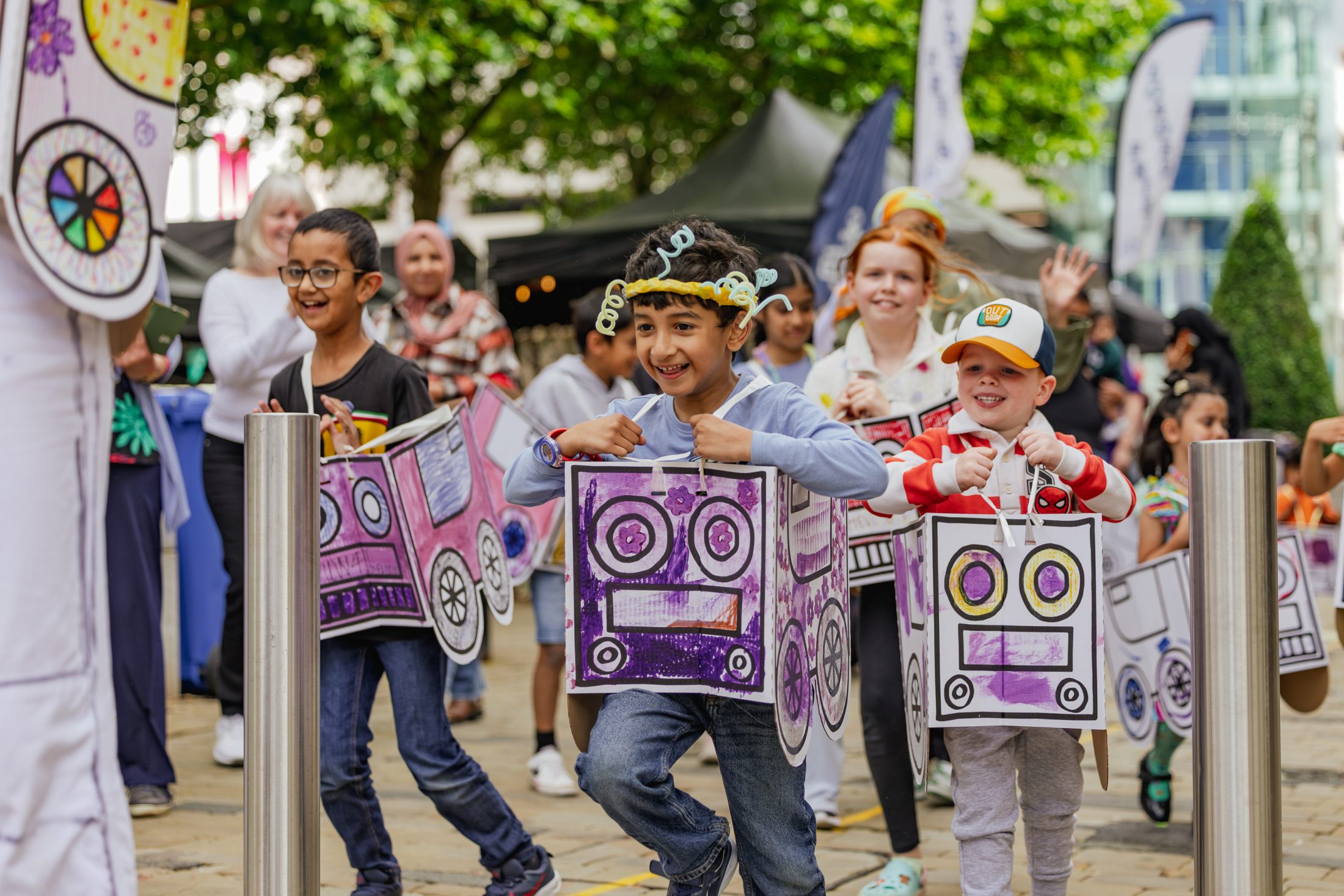 A group of children wearing colourful, homemade cardboard car costumes smile and walk in a parade outside, with adults and more people watching in the background. Trees and market stalls line the street.