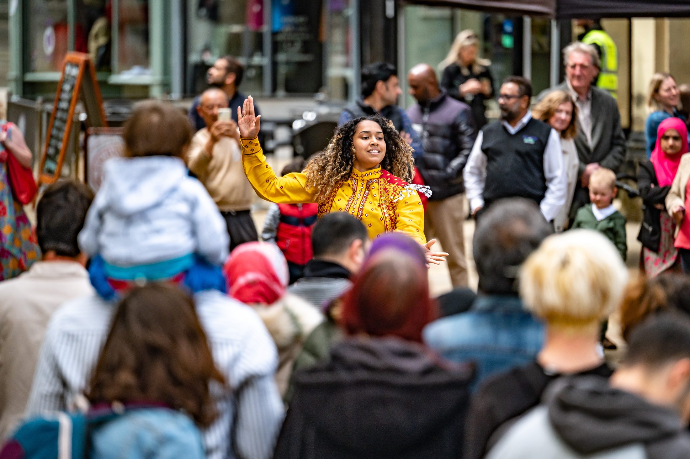 A woman in a yellow outfit dances joyfully in front of a diverse crowd outdoors. Some people watch her whilst others talk among themselves. A child sits on an adult’s shoulders, and colourful clothing fills the lively scene.