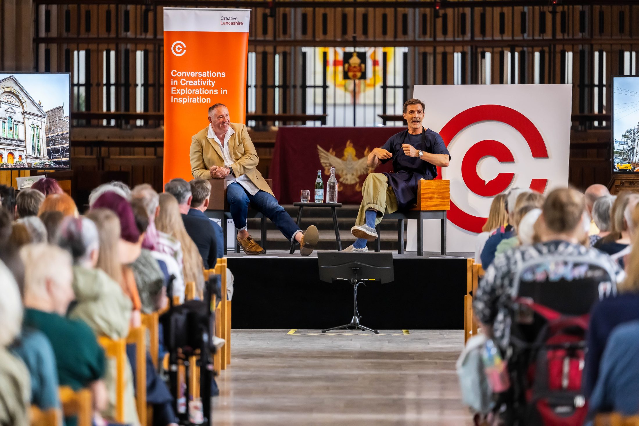 Patrick Grant and Keith Brymer Jones sit on stage in front of an audience during a live talk. Behind them are bright banners reading Conversations in Creativity Exploration Inspiration. The event is held in a large cathedral with wooden seating.