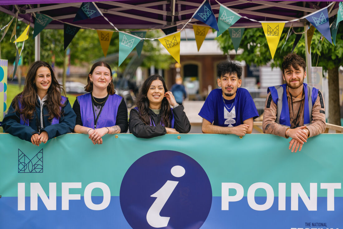 Five young adults wearing purple vests stand behind an Info Point booth outdoors, smiling at the camera. Colourful bunting decorates the tent above them, and trees are visible in the background.