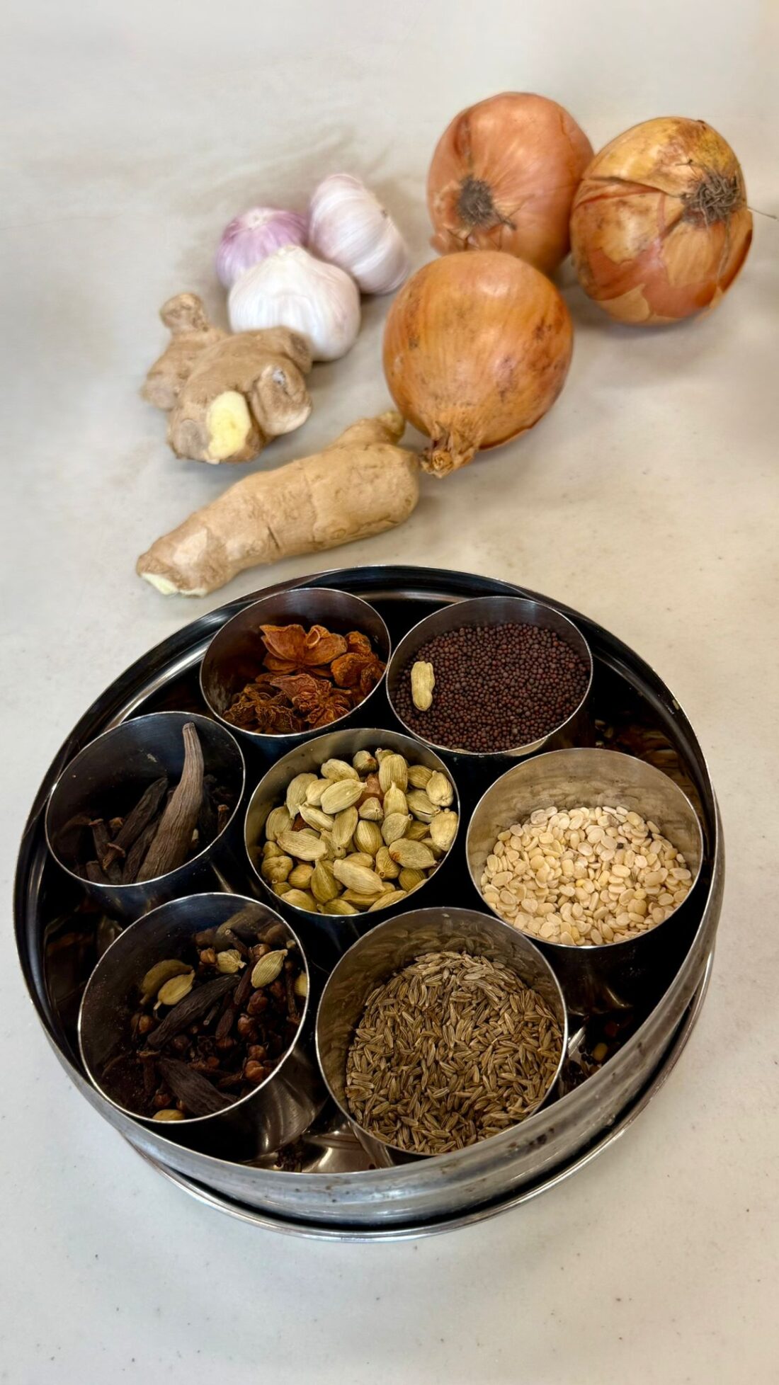 A metal spice tin with compartments holding various spices sits on a surface. Behind it are onions, garlic bulbs, and a piece of ginger root.