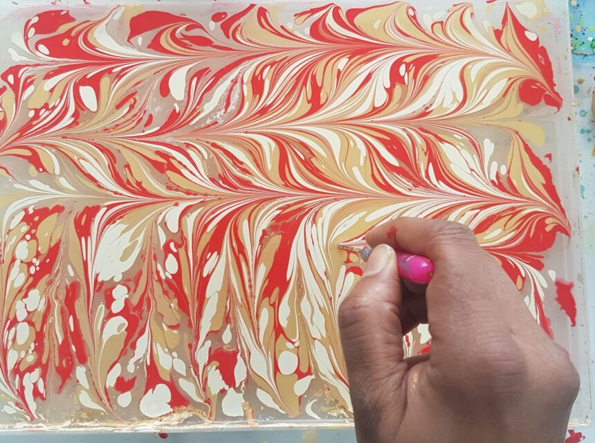 A hand uses a pink stylus to create marbled patterns with red, yellow, and white paint on a watery surface, as part of a paper marbling art technique.