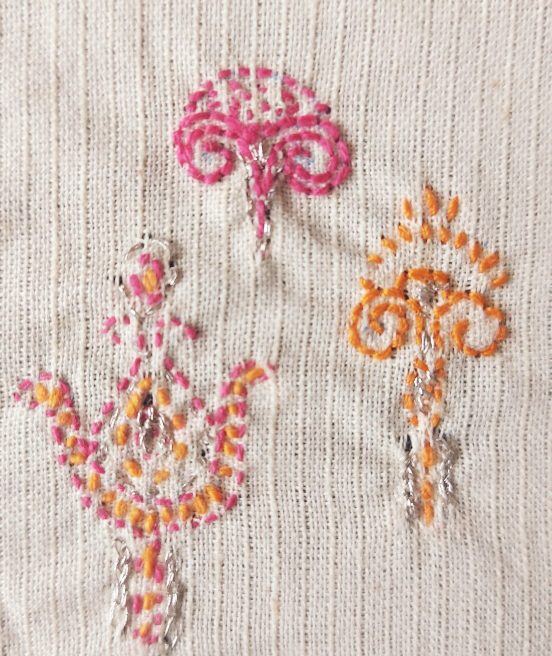 Three embroidered floral designs in pink, orange, and silver thread are stitched on a textured off-white fabric background.
