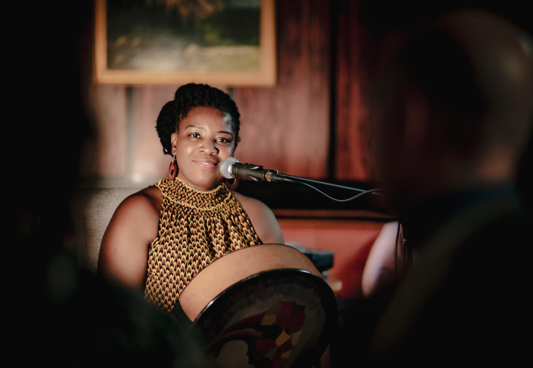 A woman in a patterned dress plays a drum and smiles whilst sitting at a microphone in a warmly lit room, with blurred figures in the foreground.