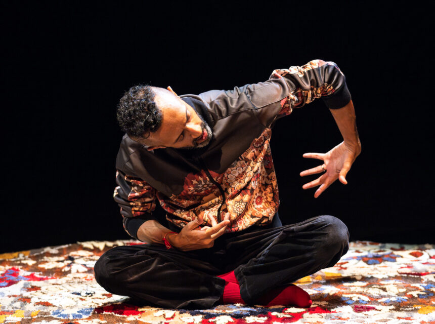 A person with short curly hair sits cross-legged on a colourful rug, wearing a patterned jacket and red socks, performing an expressive hand gesture against a dark background.