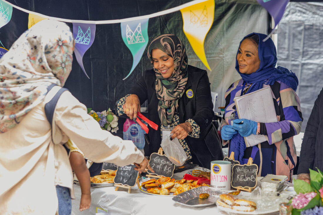 Two women in hijabs serve baked goods at a decorated outdoor stall, smiling at a customer. Colourful flags hang above, and various pastries are displayed on the table with handwritten signs.