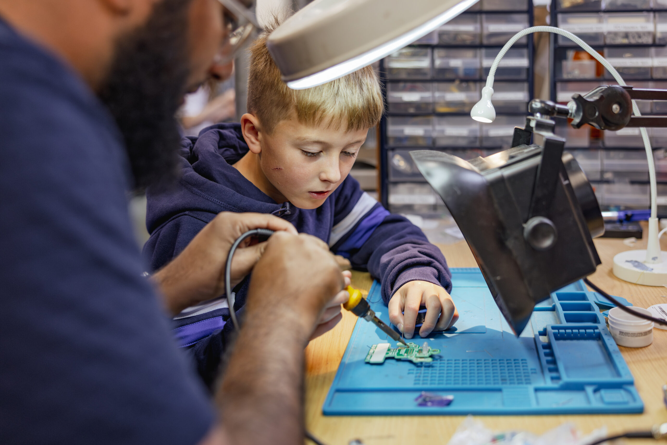 Child watches adult soldering a circuit board under a lamp, surrounded by tools, in a workshop setting.