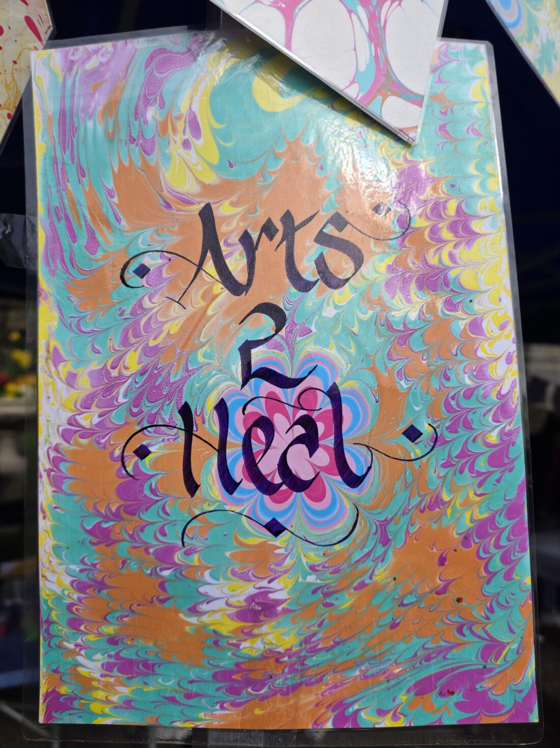 A colourful, marble-patterned sign with the words Arts 2 Heal in black calligraphy, surrounded by swirling, vibrant colours like orange, teal, pink, yellow, and blue.