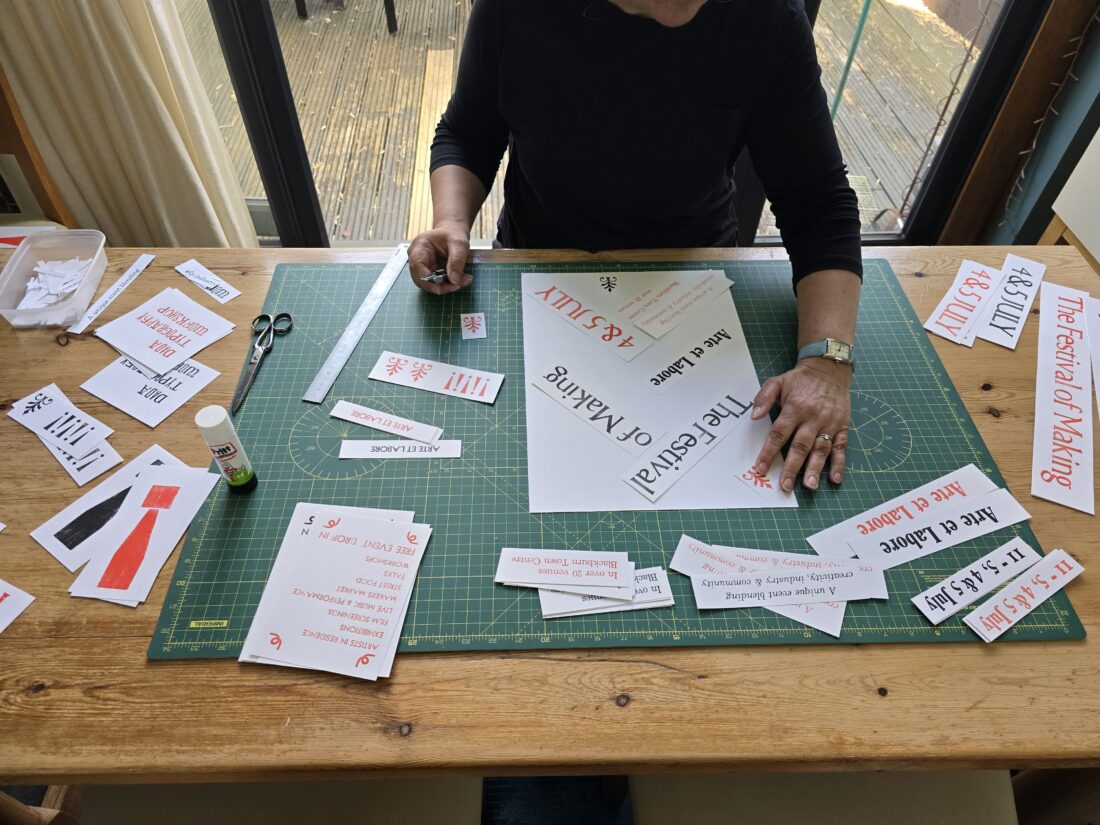 A person arranges printed event signs on a cutting mat, surrounded by paper scraps, scissors, and glue on a wooden table near glass doors.