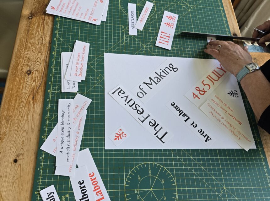 A person is cutting paper on a green cutting mat with various printed phrases like The Festival of Making and Arte et Labore scattered around. A glue stick and metal ruler are also on the wooden table.