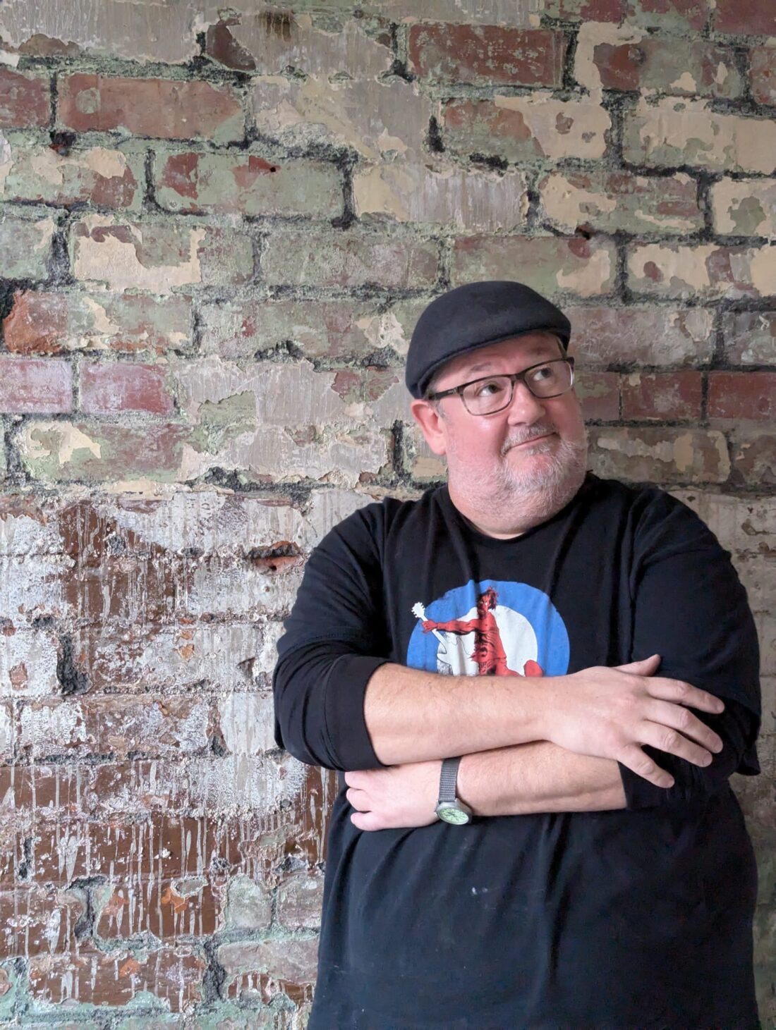 A man wearing glasses, a black cap, and a black shirt with a graphic stands with arms crossed, leaning against a weathered brick wall, looking slightly upwards and to the right.