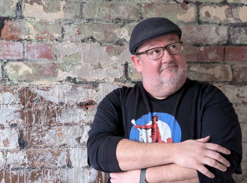A man wearing glasses, a black cap, and a black shirt with a graphic stands with arms crossed, leaning against a weathered brick wall, looking slightly upwards and to the right.