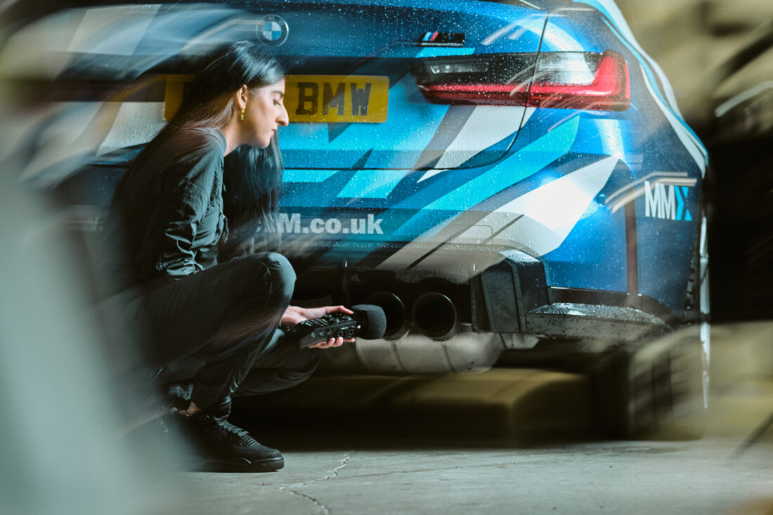 A woman crouches by the exhaust of a blue BMW sports car, holding a device, possibly measuring sound or emissions, in an indoor garage setting.