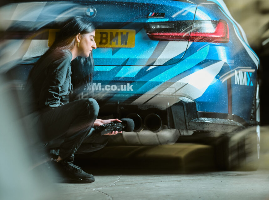 A woman crouches by the exhaust of a blue BMW sports car, holding a device, possibly measuring sound or emissions, in an indoor garage setting.