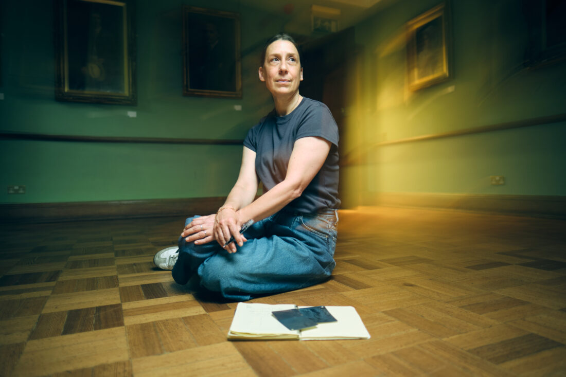 A person sits cross-legged on a wooden floor in a gallery, with a notebook and mobile in front of them. Framed portraits hang on green walls in the background.