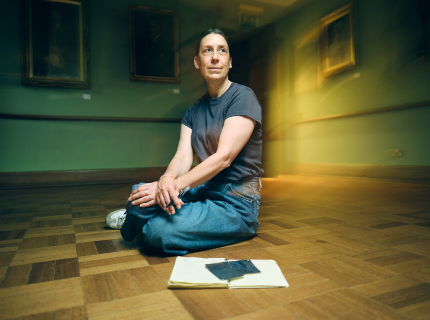 A person sits cross-legged on a wooden floor in a gallery, with a notebook and mobile in front of them. Framed portraits hang on green walls in the background.