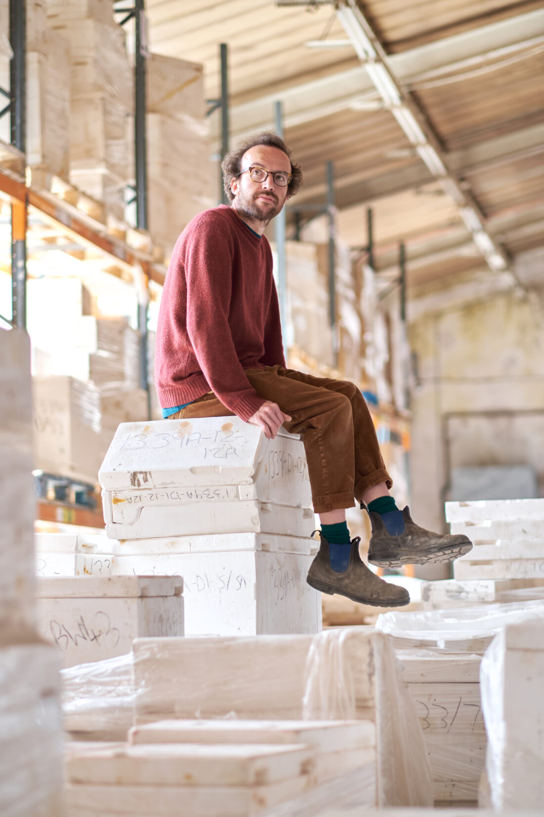 A person wearing glasses, a red jumper, brown trousers, and boots sits on stacked white boxes in a warehouse with shelves and a high ceiling.
