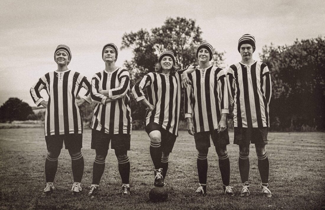 Five people in vintage football kits and hats stand in a line on a pitch, arms on hips or folded, posing confidently. The photo is black-and-white, giving an old-fashioned feel.