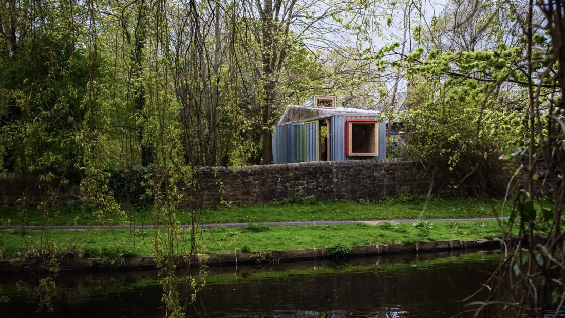A small, colourful shed stands on a stone wall, surrounded by trees and greenery, near a grassy area and a calm river in the foreground.