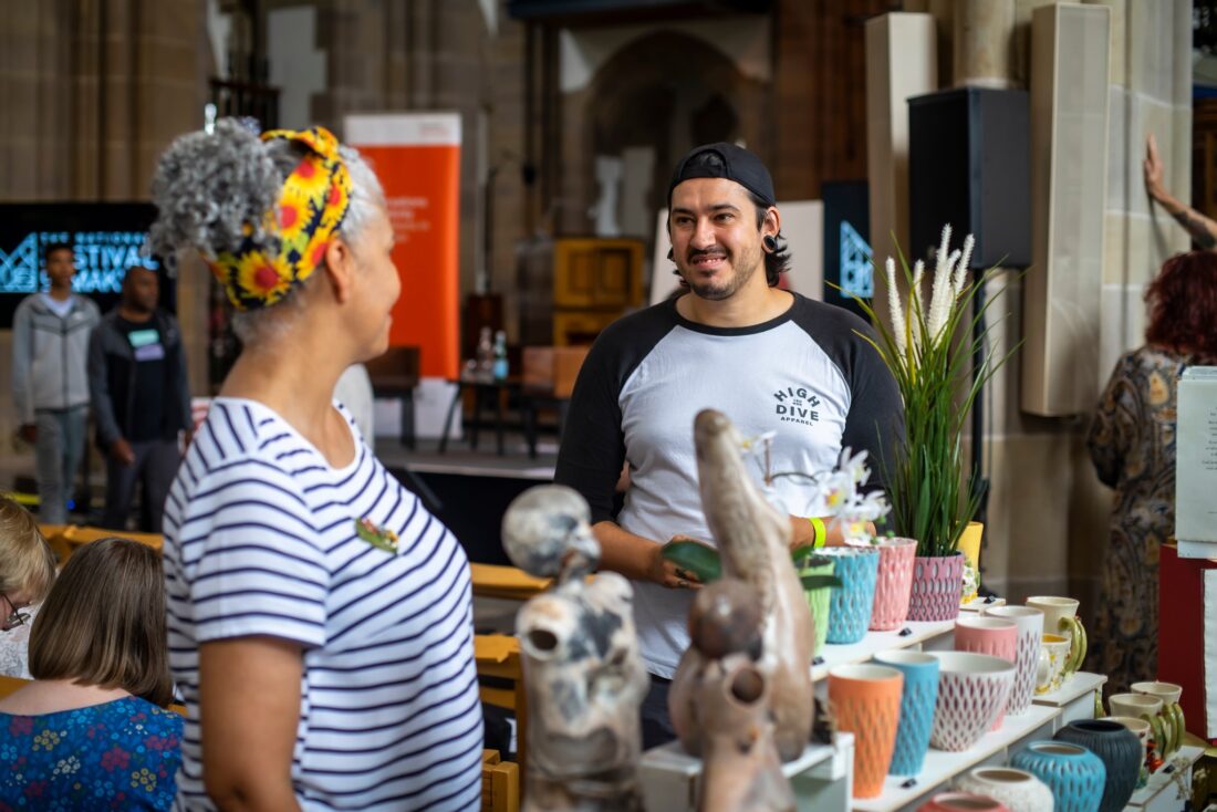 A woman and man smile and chat at a pottery stall indoors, surrounded by ceramic art and colourful cups, with other people and displays in the background.