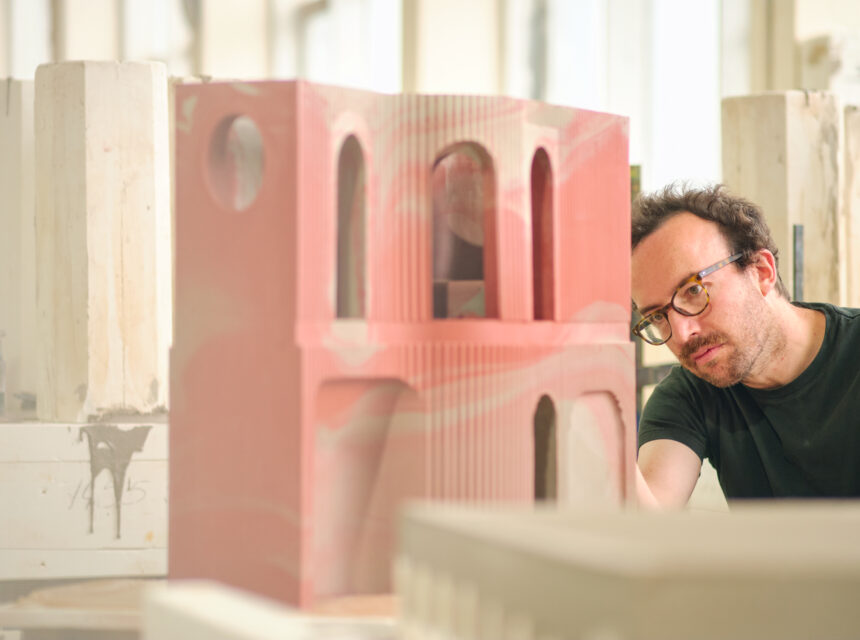 A man with glasses closely examines an architectural model made of pink material, surrounded by other model structures in a bright room.