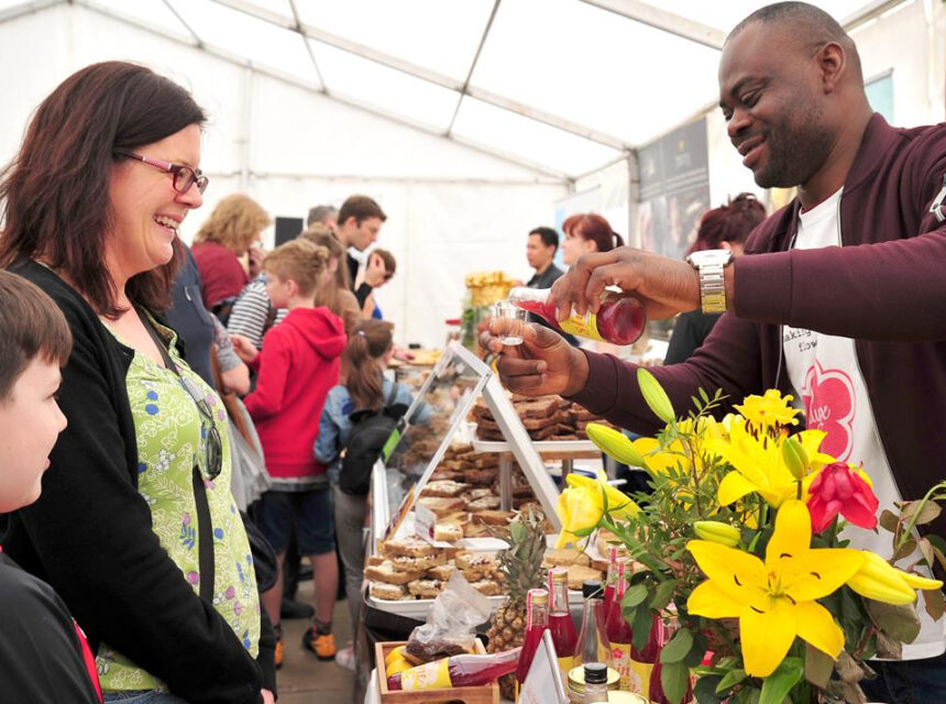 A man with a maroon jacket pours a sample of red liquid from a bottle into a small glass for a woman and a boy at a food festival stall. In the foreground, a vibrant bouquet of yellow lilies sits next to various baked goods and products.