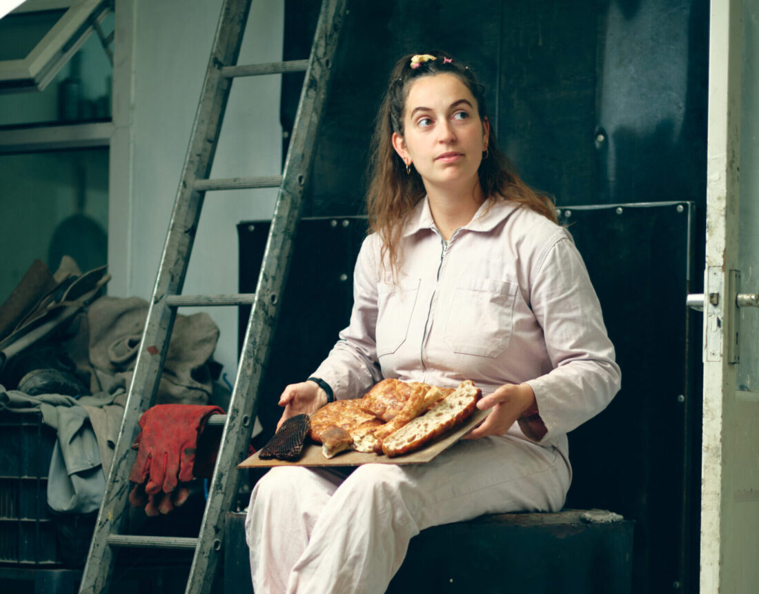 A woman in a light jumpsuit sits by a ladder, holding a tray of baked bread and pastries, looking off to the side.