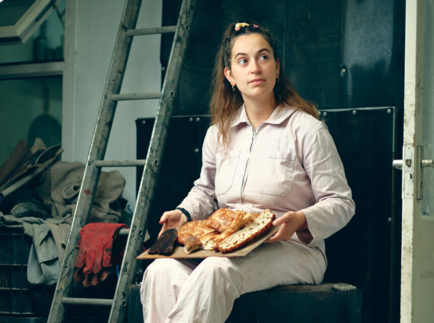 A woman in a light jumpsuit sits by a ladder, holding a tray of baked bread and pastries, looking off to the side.
