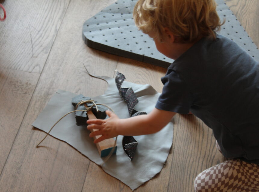 A young child reaches for pieces of fabric, string, and leather laid out on a wooden floor, appearing to work on a craft or art project.