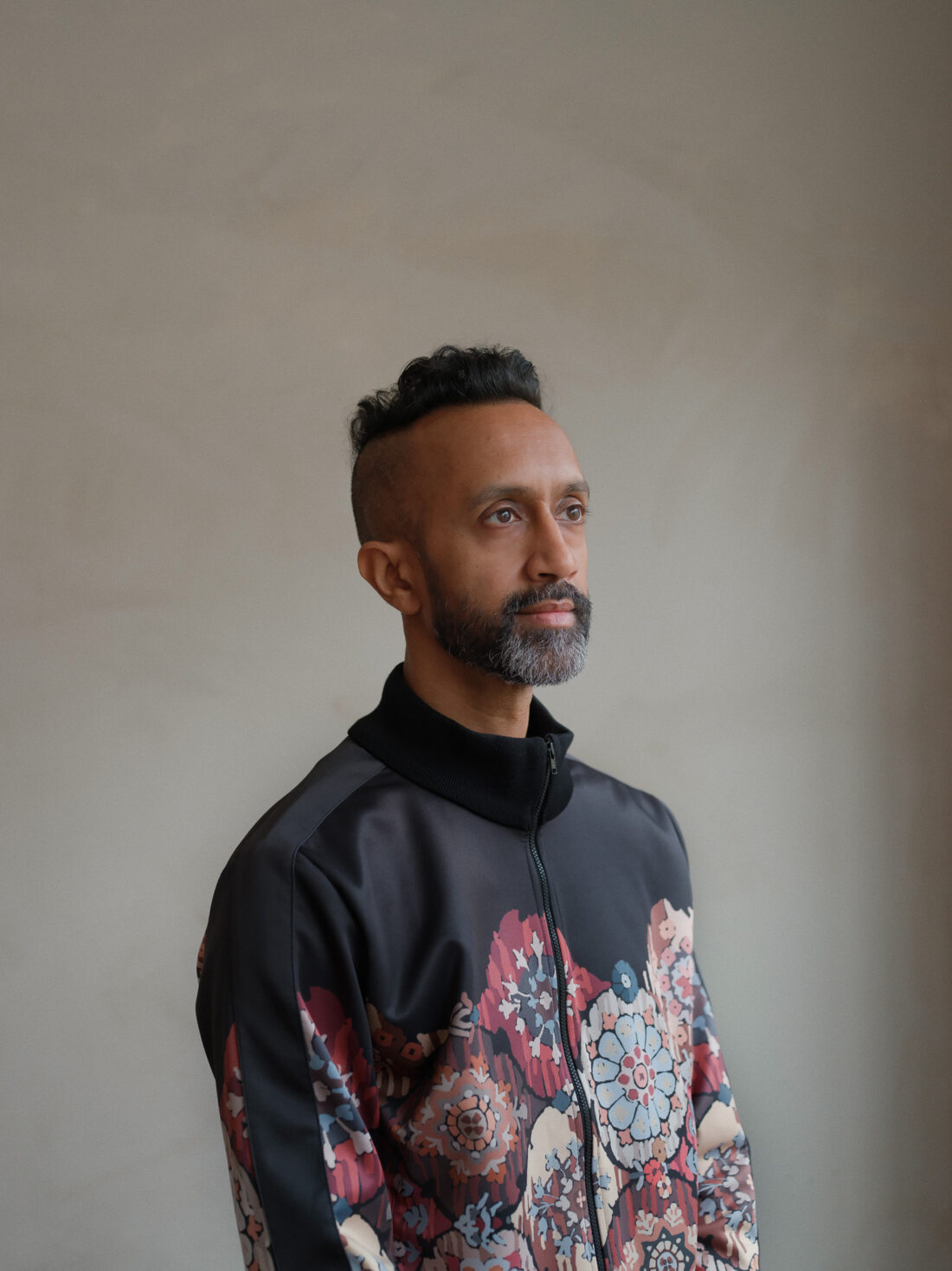 A man with short curly hair and a beard wears a dark, floral-patterned jacket, standing against a plain beige wall whilst gazing thoughtfully to the side.