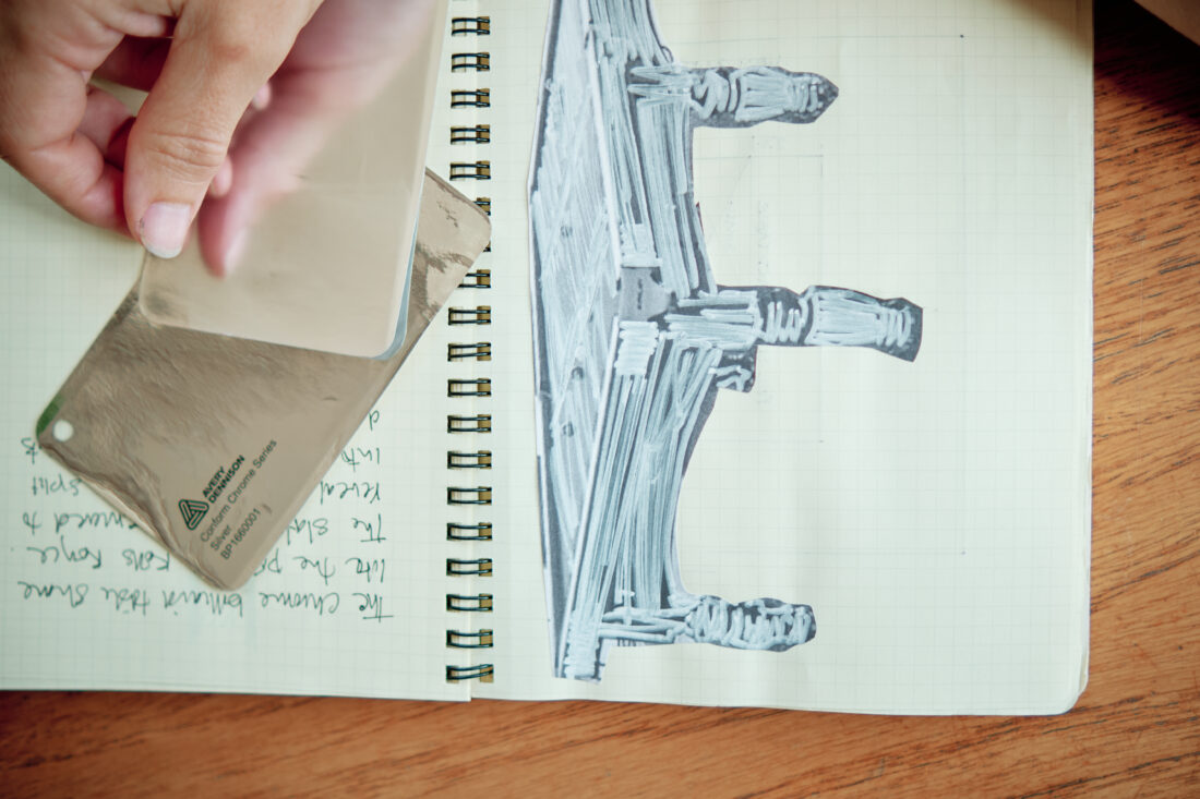 A hand holds a metallic colour sample card above a sketch of architectural columns in a spiral notebook with handwritten notes, on a wooden table.