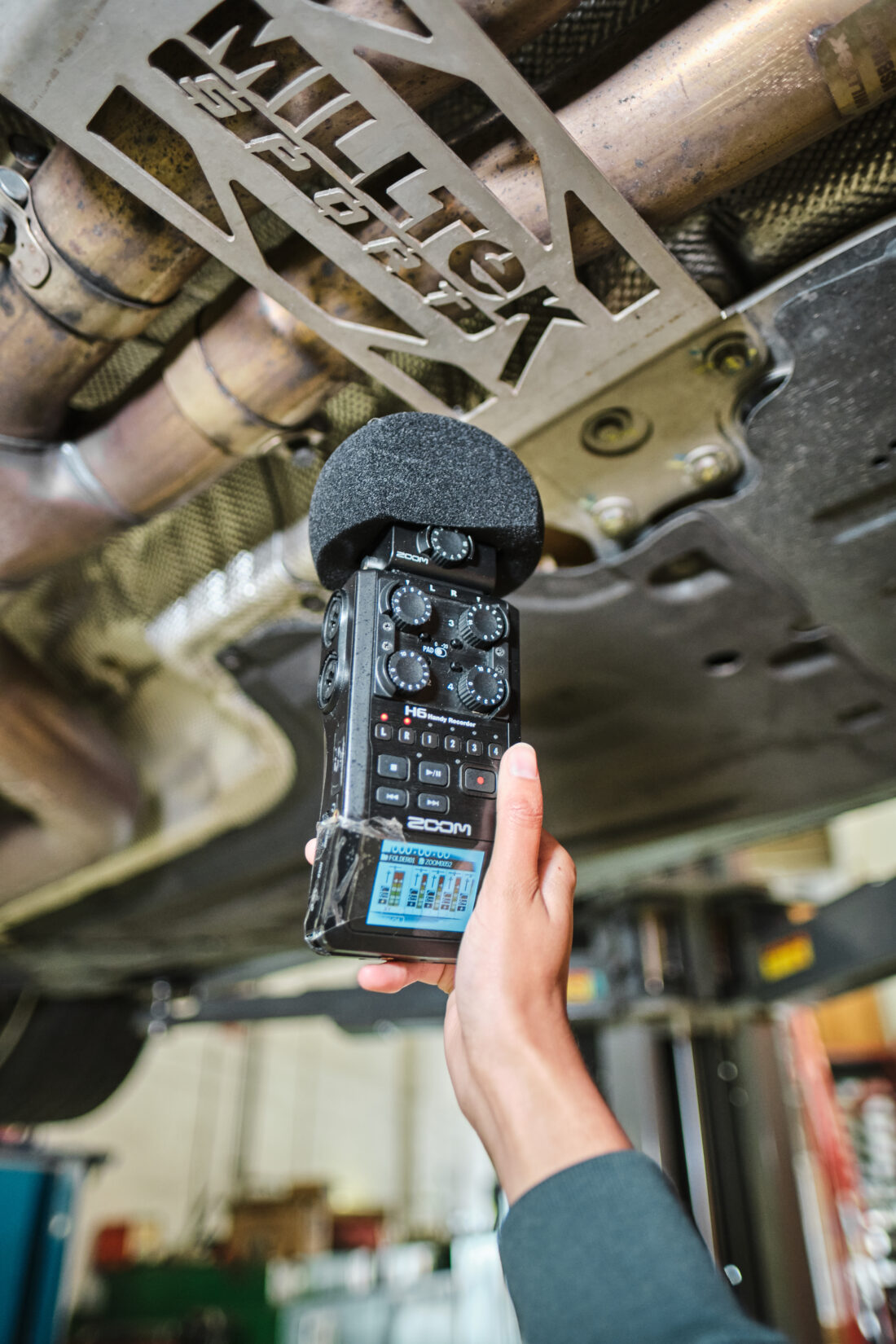 A hand holds a portable audio recorder with a microphone near the exhaust system of a car lifted on a ramp. Milltek Sport is visible on a metal car part above.