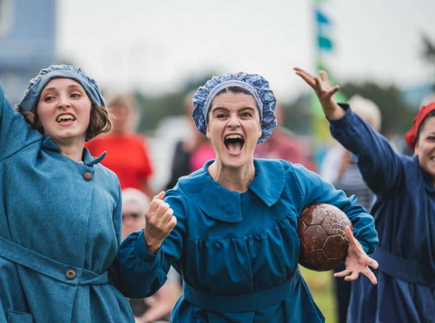 Three women in blue historical costumes and bonnets enthusiastically cheer whilst holding a vintage leather football outdoors, with people and greenery blurred in the background.